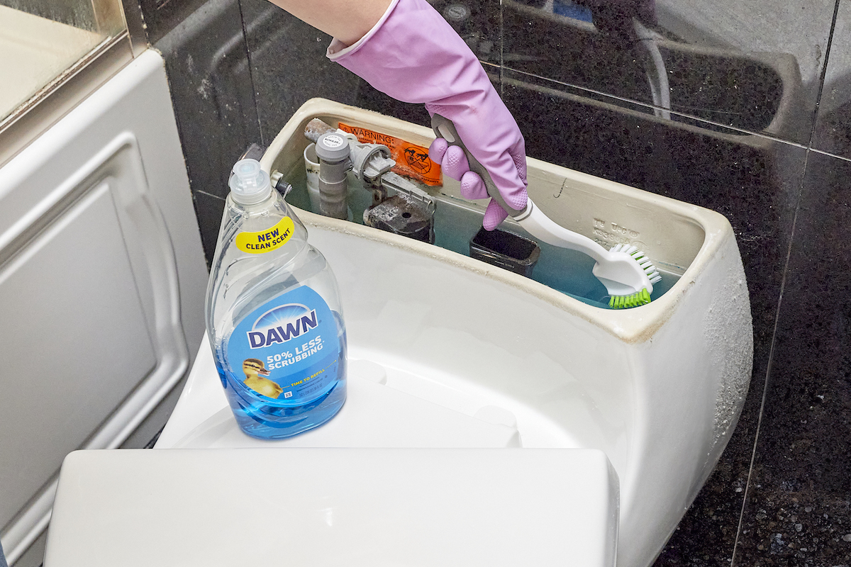 Woman cleans inside of a toilet tank with a scrub brush, with a bottle of Dawn dish soap on the seat.