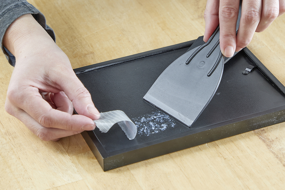 Woman uses a plastic putty knife to remove duct tape from the back side of a black picture frame.