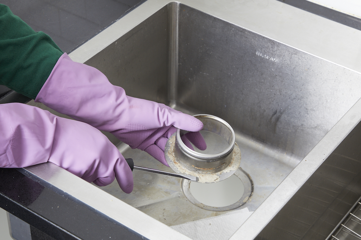 Person wearing rubber gloves removes old plumber's putty with a screwdriver from the underside of a kitchen sink flange.