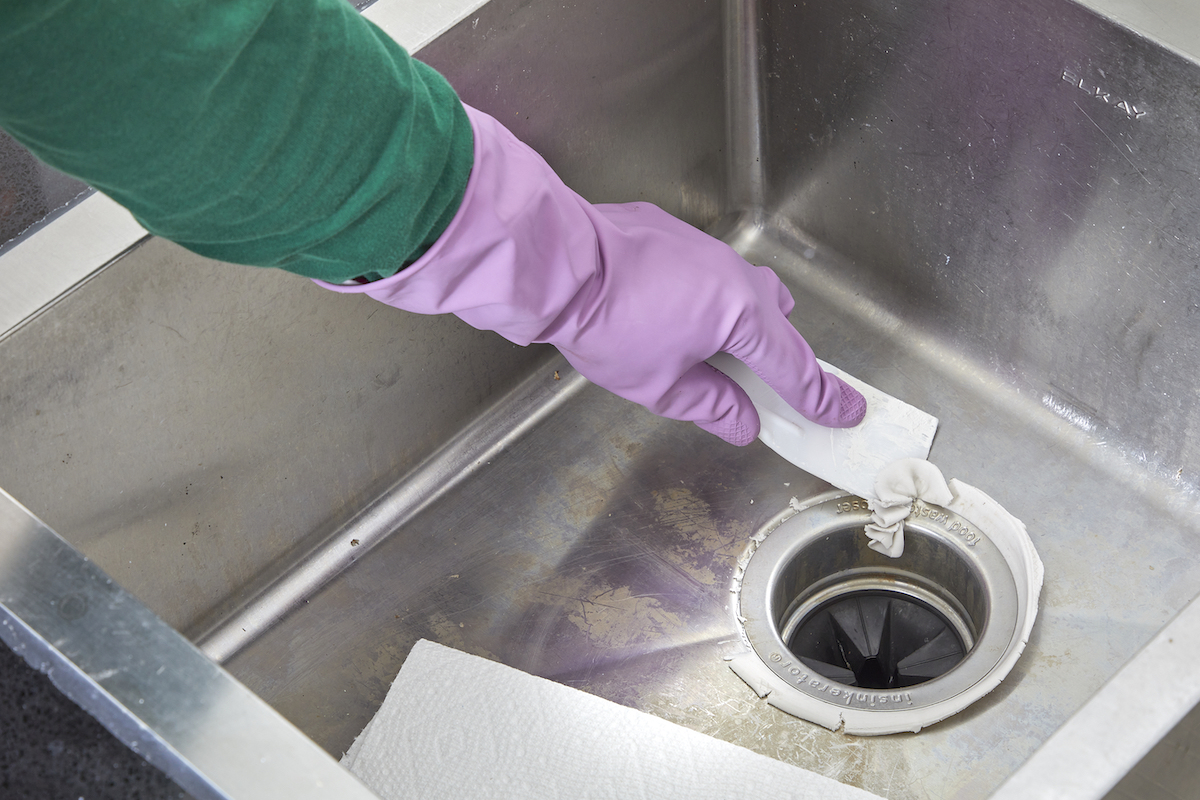 Woman wearing rubber gloves scrapes excess fresh plumber's putty from around the sink flange of a garbage disposal.