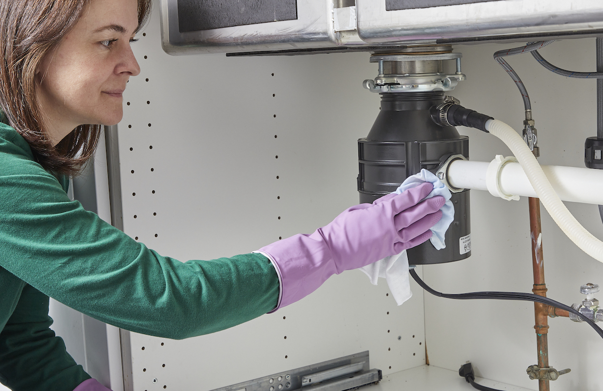 Woman wearing rubber gloves wipes a leak from the pipe leading into the side of a garbage disposal under the kitchen sink.