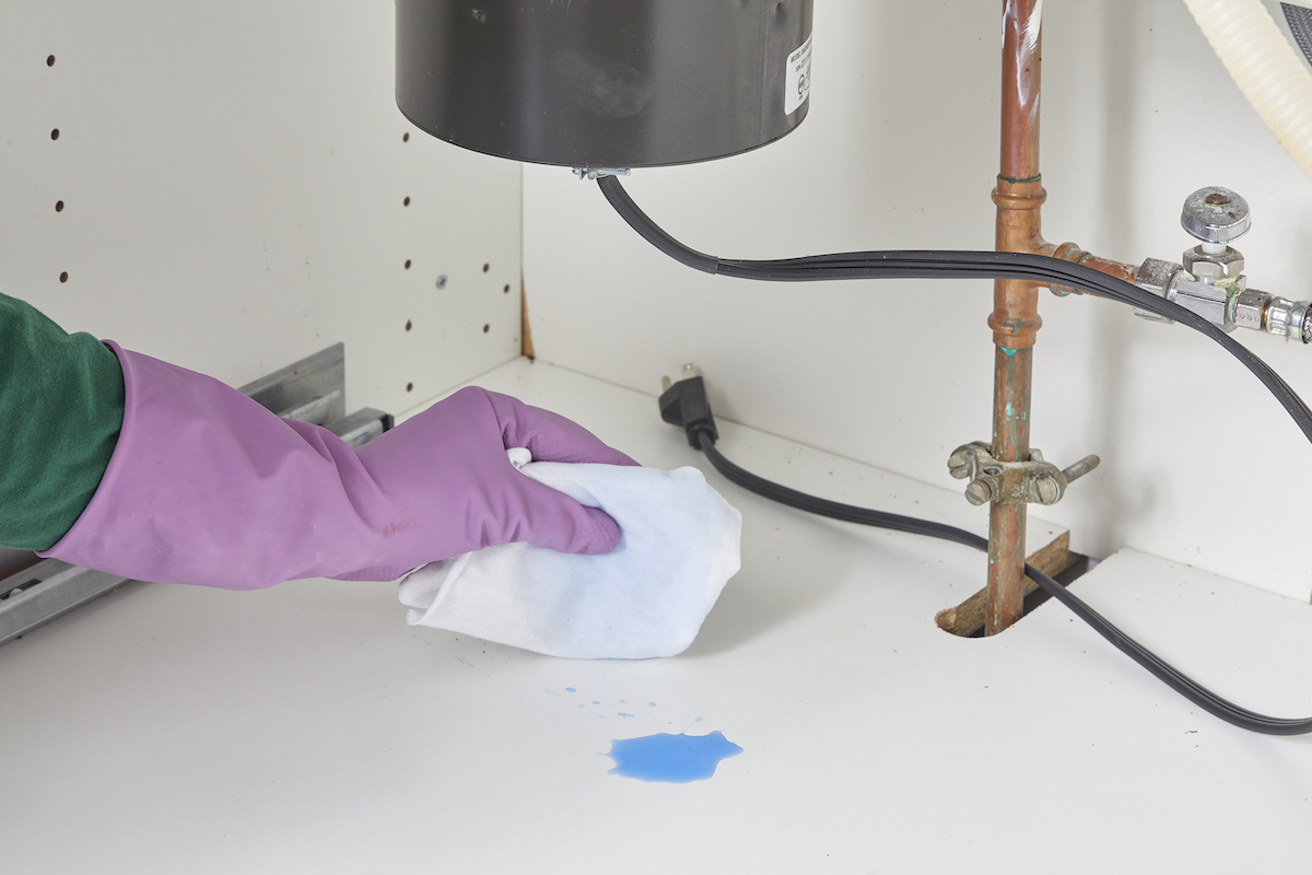 Person wearing rubber gloves wipes drops of dyed blue water from cabinet under a garbage disposal.