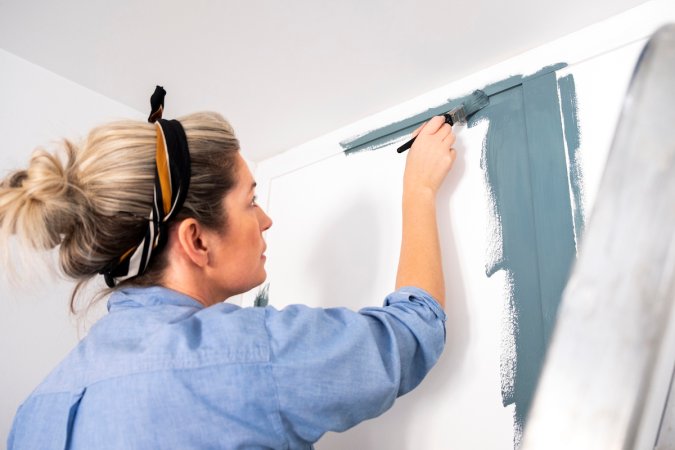 A woman wearing casual clothing and a headband is painting her wood panelled wall with blue paint while standing on a ladder.