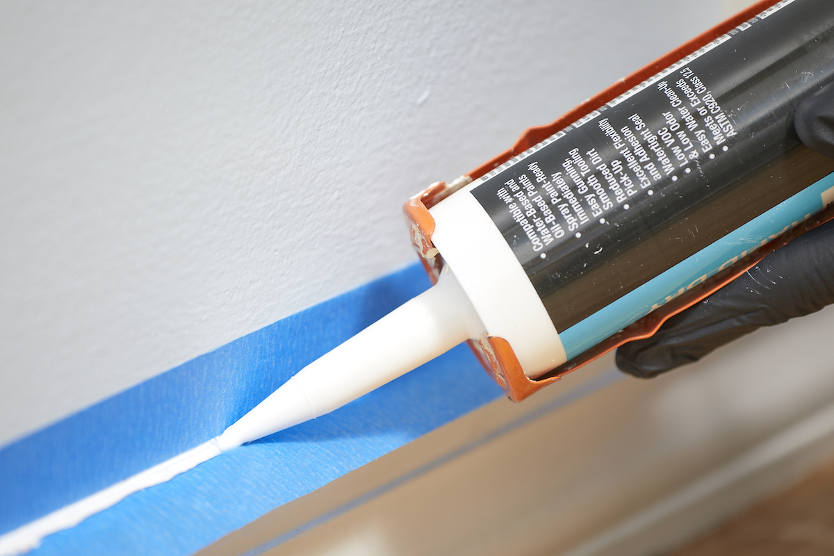 Close up view of caulk gun caulking a baseboard over painter's tape.