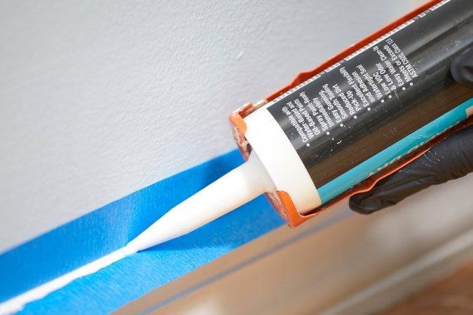 Close up view of caulk gun caulking a baseboard over painter's tape.