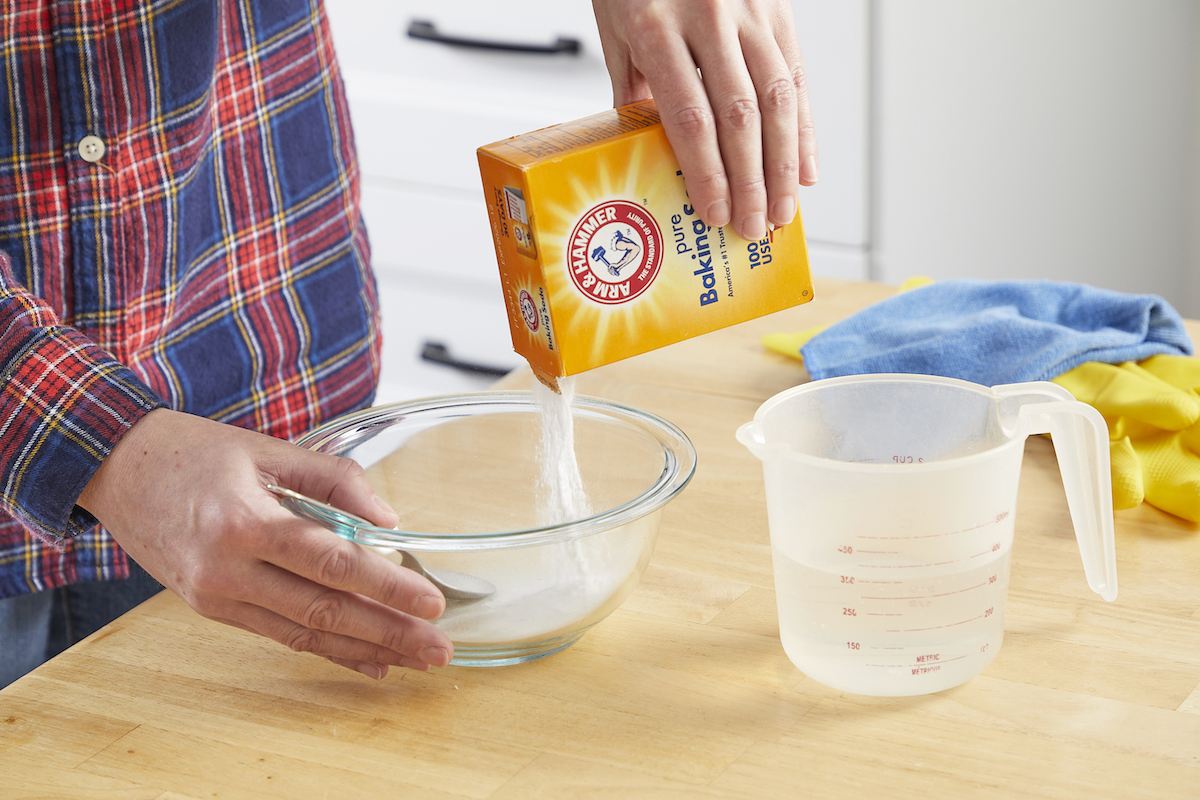 Woman wearing a plaid shirt mixes baking soda and water in a glass bowl.