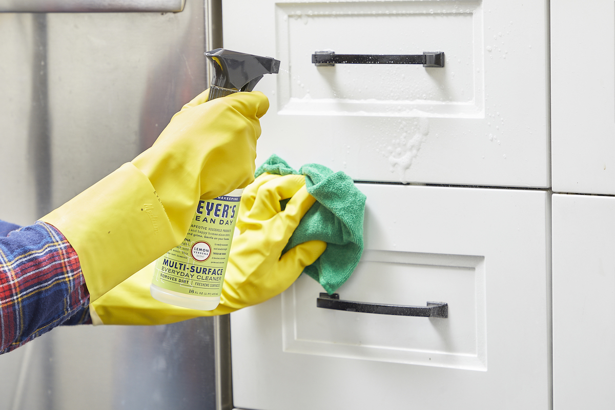 Woman wearing yellow cleaning gloves sprays citrus cleaner on white cabinets.