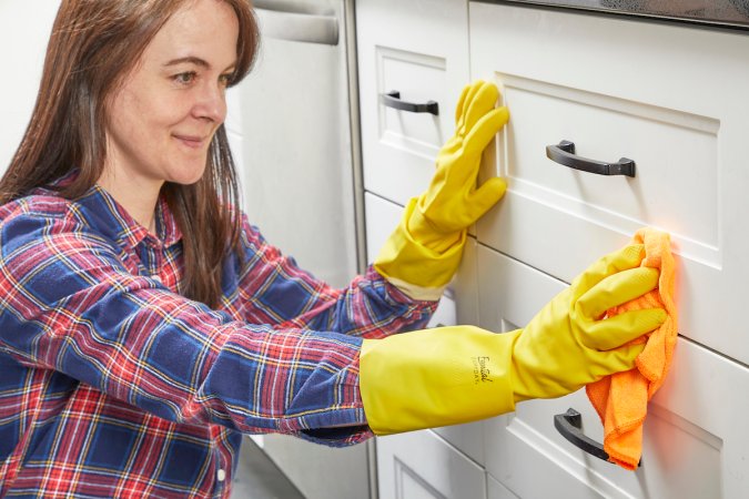 Woman wearing a plaid shirt and yellow cleaning gloves wipes white kitchen cabinets with a cloth.