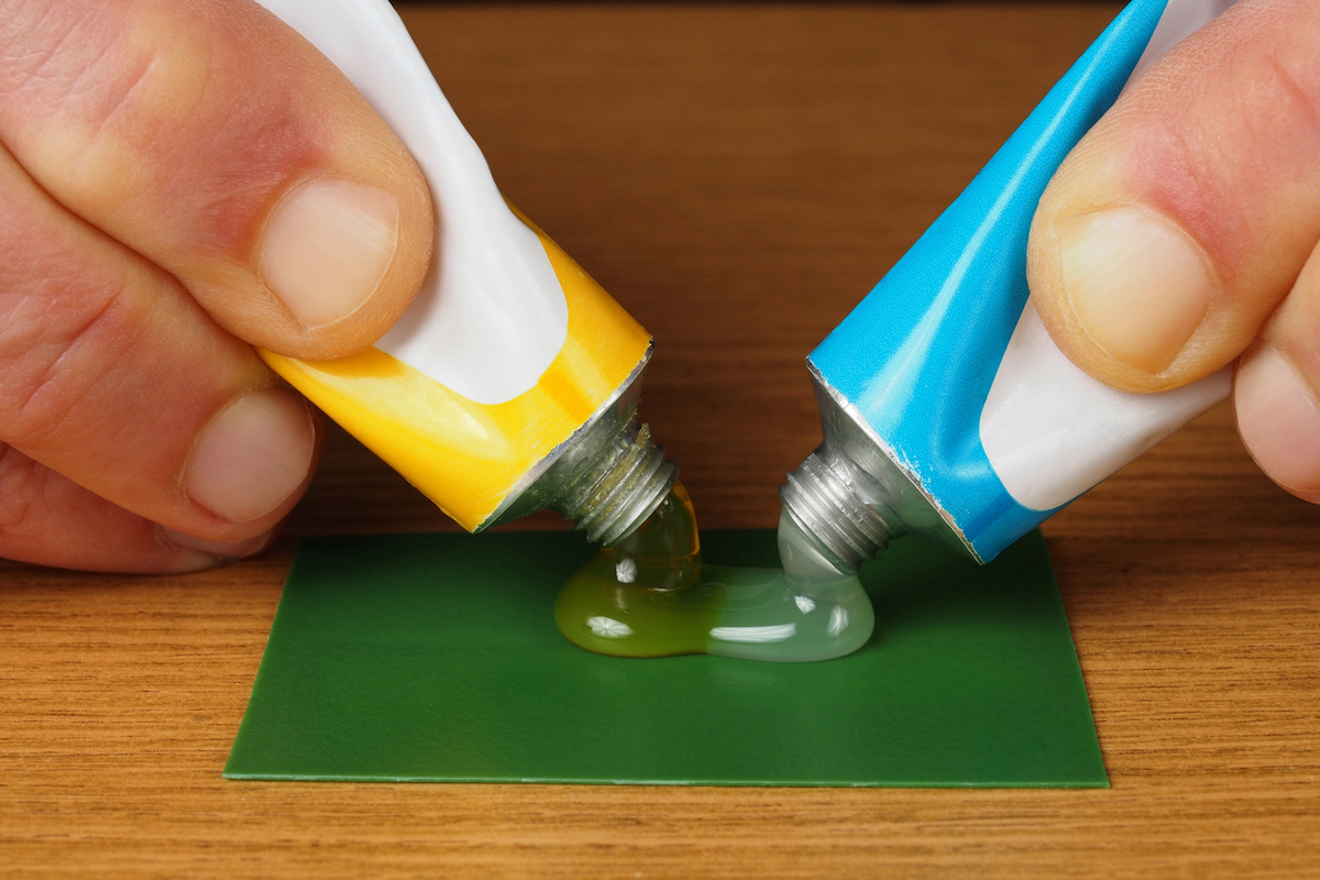 A person is squeezing a tube of epoxy glue in each hand so they mix together on a green square.