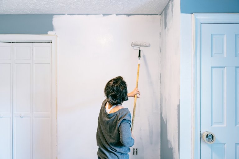 A woman with a grey sweater uses a tall roller brush to paint an interior wall with white paint.