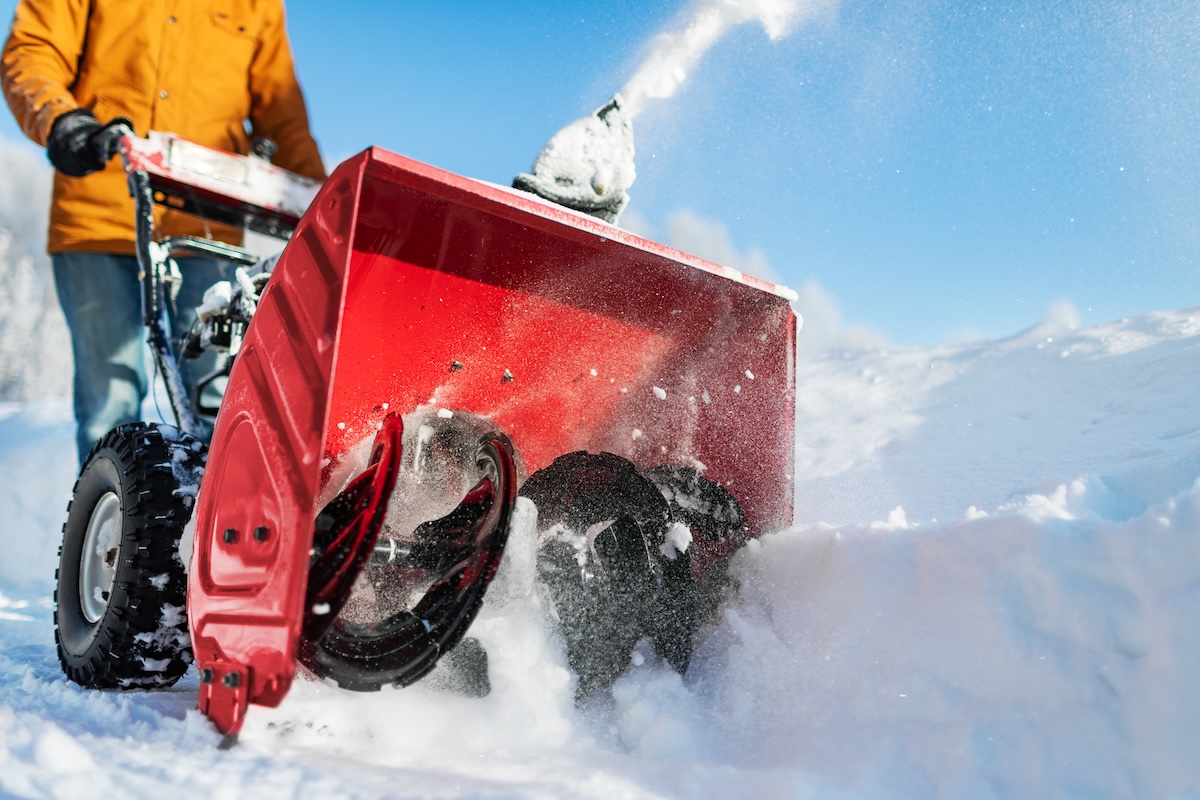 A man in a yellow coat pushes a snow blower on a sunny day.