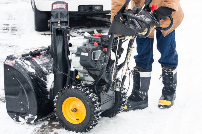 A man in a brown coat works to fix a snowblower on a snowy day.