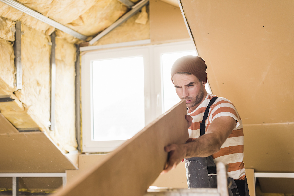 Young man checking plank on construction site under dormer window with exposed insulation.