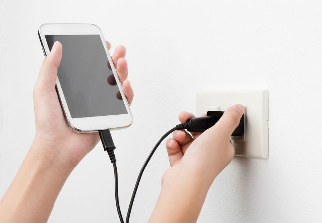 A close up of a person plugging in a smartphone into an outlet in a white wall.