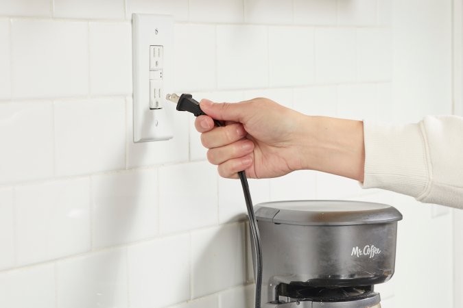 Woman plugs coffee maker into an electrical outlet in a kitchen.