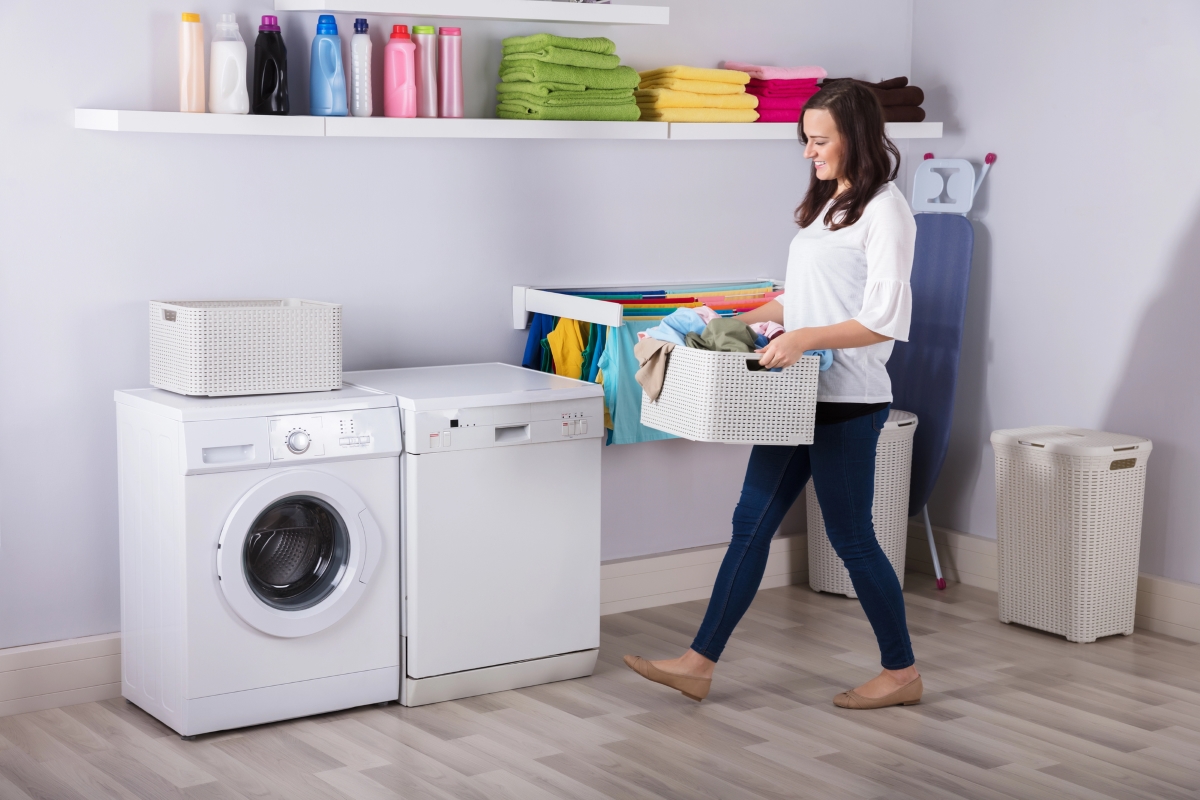 A woman is walking towards the washer and dryer with a basket of clothes.