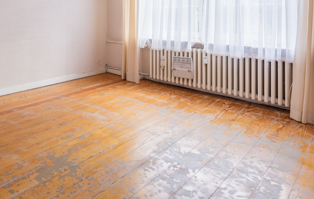 The hardwood floor of an empty bedroom with curtains in the window is damaged with deep scratches.