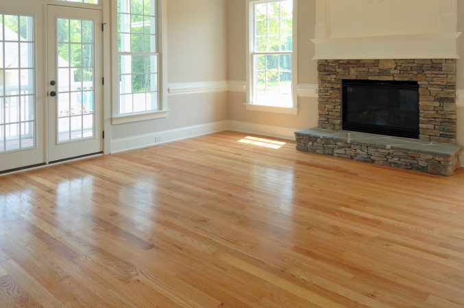 Freshly waxed hardwood flooring shines in the light of large windows in an empty living room.