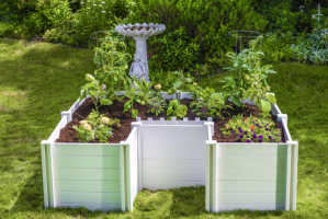 A keyhole garden made of white vinyl panels is growing a kitchen garden in front of a stone bird bath.