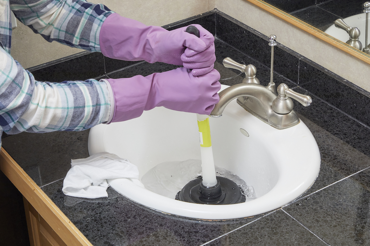 Woman wearing rubber cleaning gloves uses a plunger to plunge a bathroom sink.