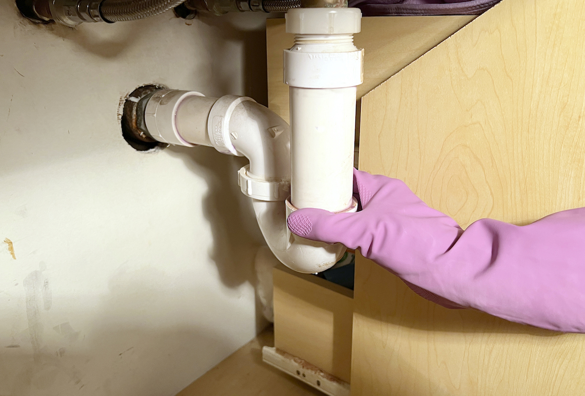 Person wearing rubber gloves touches a P-trap under the bathroom sink.