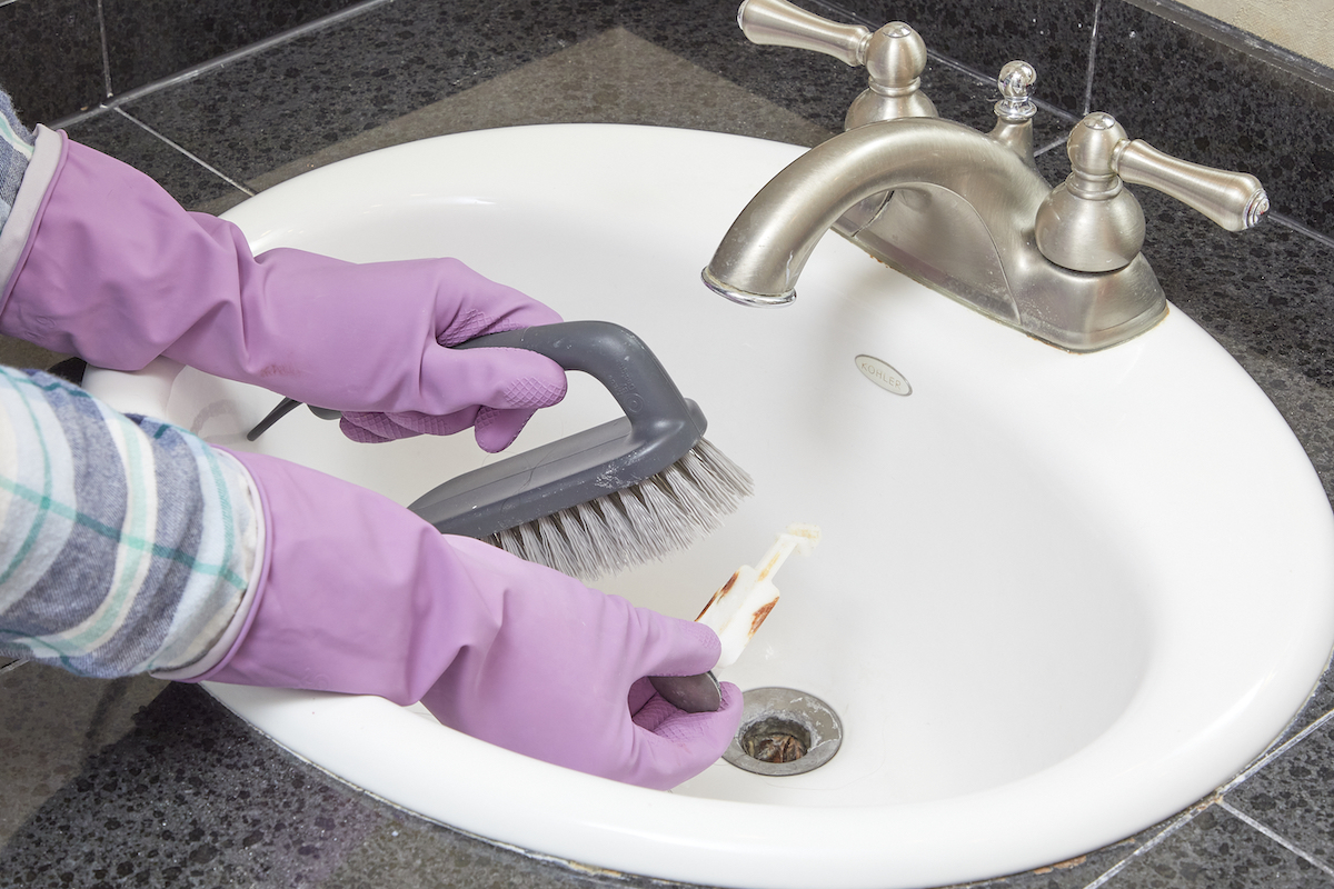 Woman wearing rubber gloves uses scrub brush to clean a sink stopper.