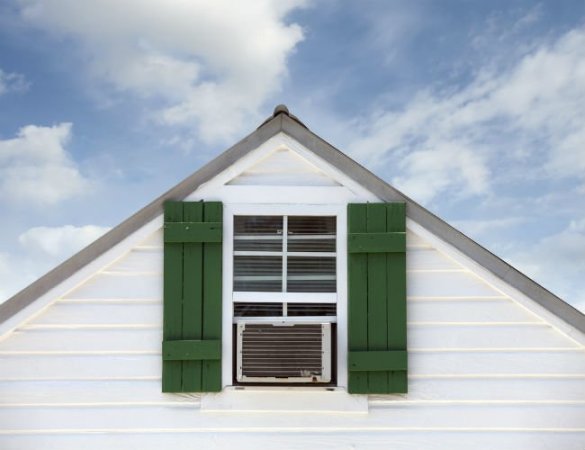 A window AC unit sits in an attic-level window on a house with white siding and green shutters.