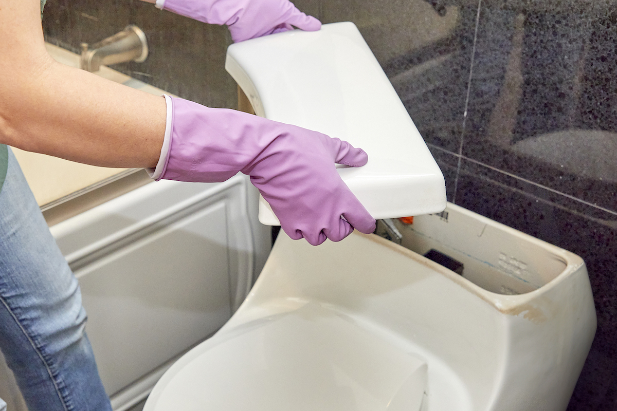 Woman wearing rubber gloves removes lid from toilet tank.