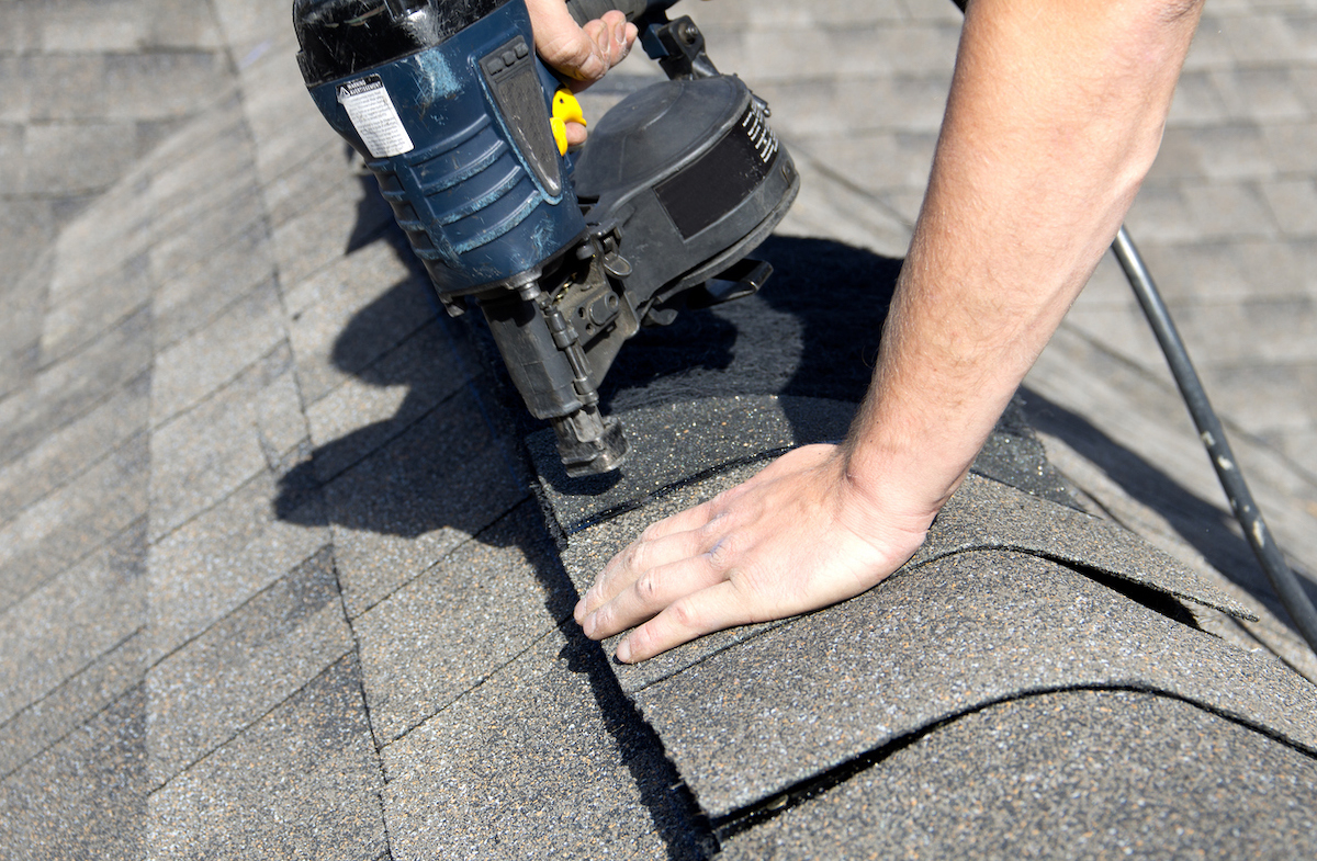 A person is nailing shingles on the peak of a house's roof.