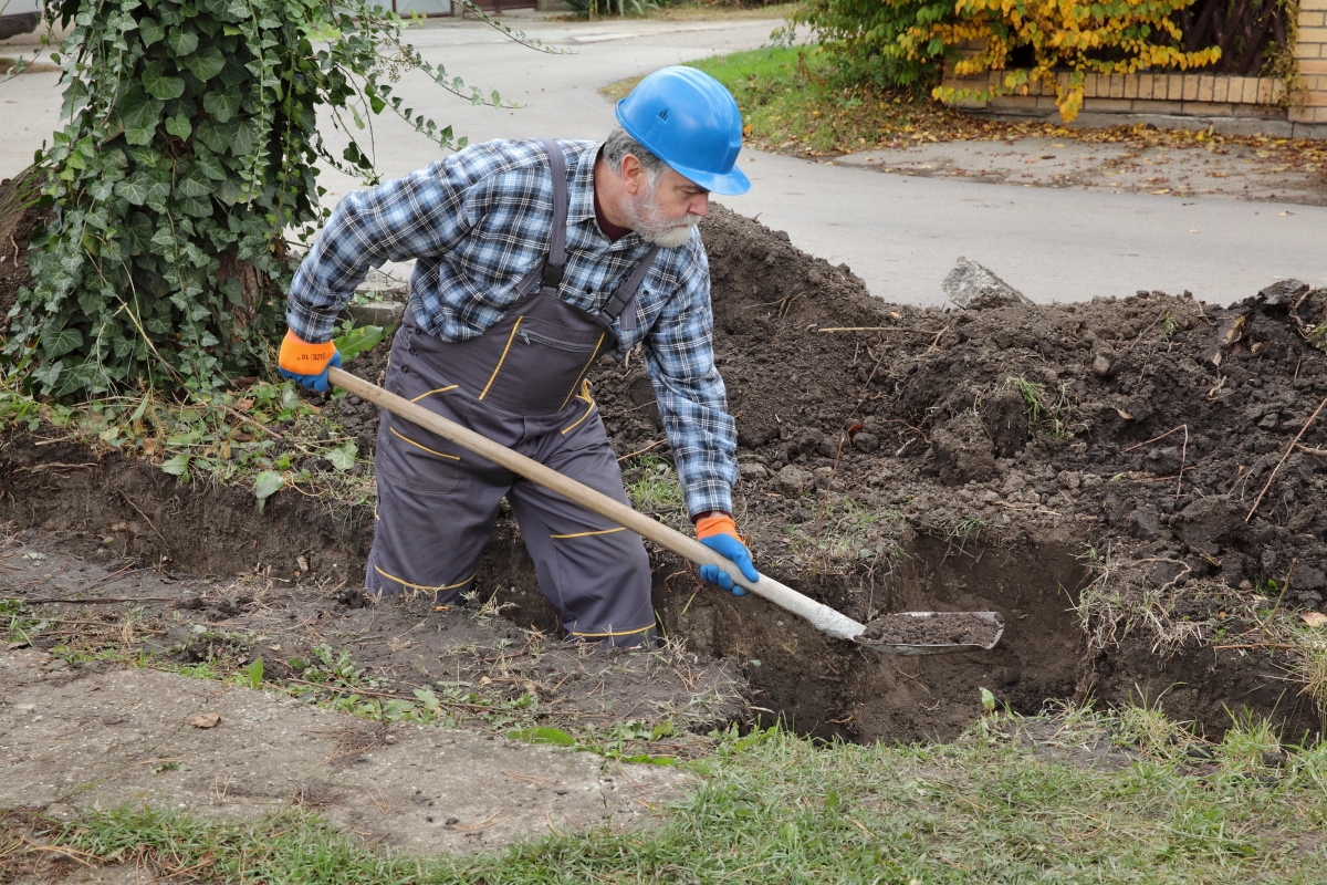 A man in a blue hard hat is digging a trench in his yard.