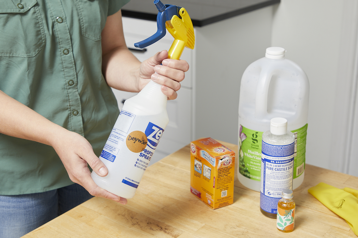 Woman holds a spray bottle labeled "degreaser" with vinegar and baking soda containers nearby.