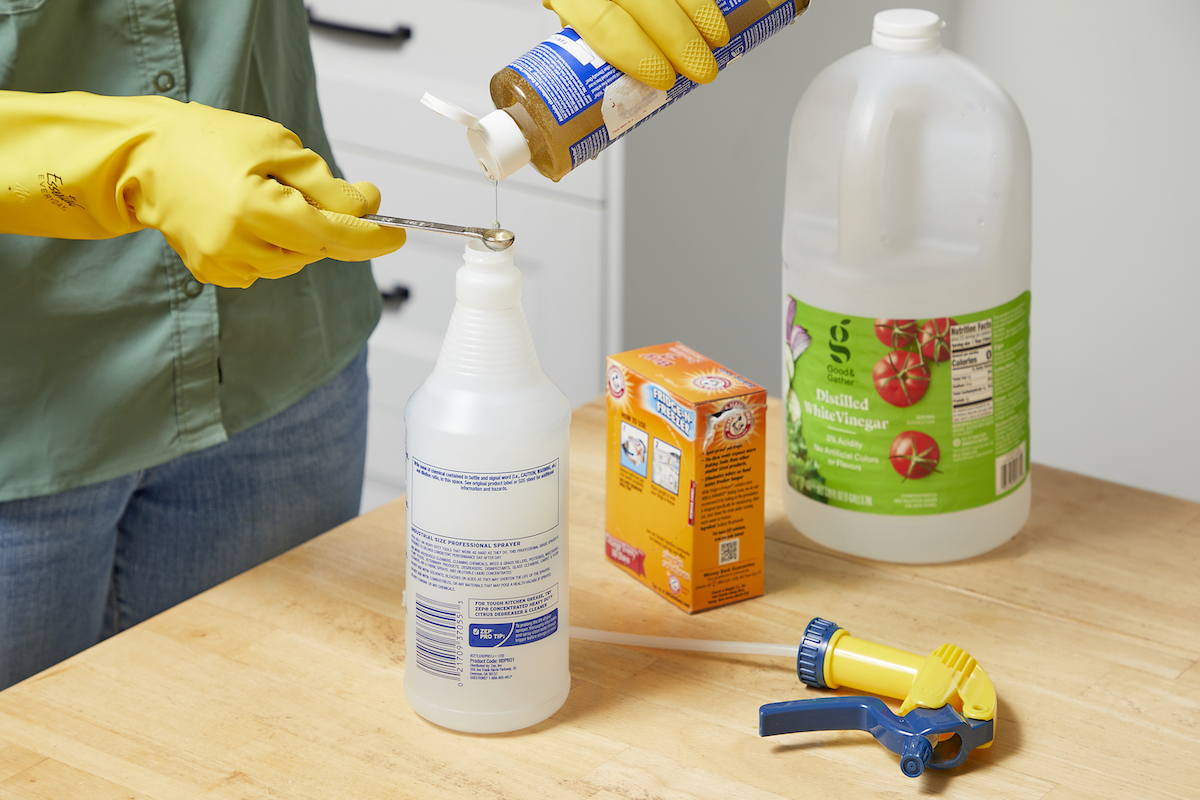 Woman measures castile soap into a spray bottle, with baking soda and vinegar containers in the background.