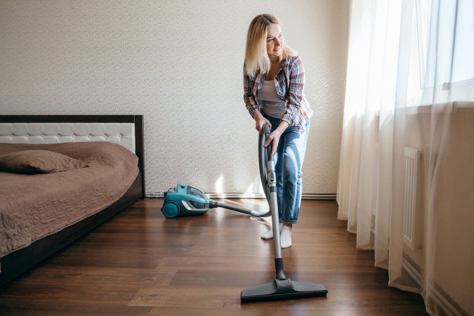 A woman is cleaning a bedroom's dark wooden laminate floor with a vacuum cleaner.