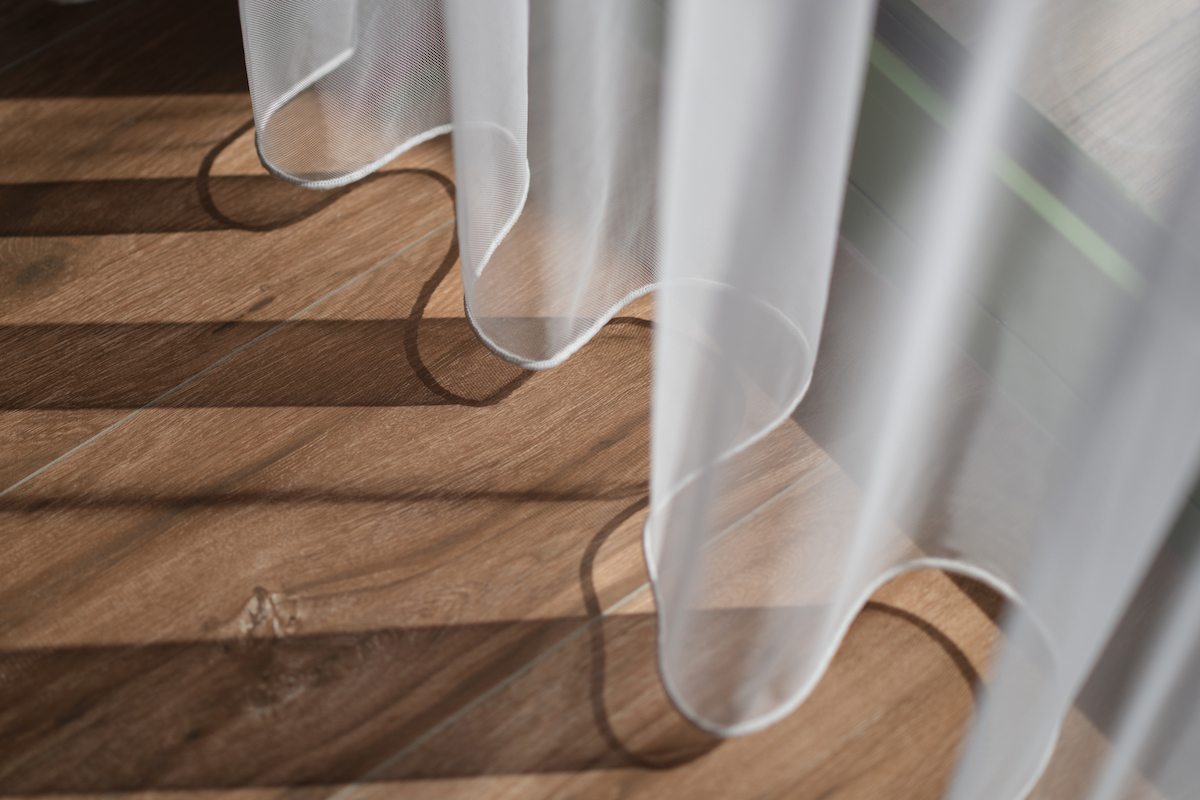 Shadows from white sheer curtains and sunlight are cast across a wooden laminate floor.