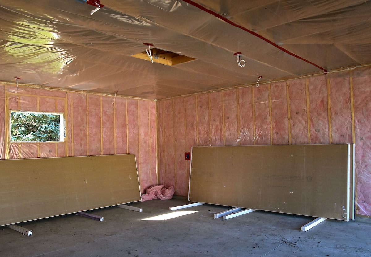 A garage is insulated with pink batt insulation; a couple of panels of drywall lean against the walls. 