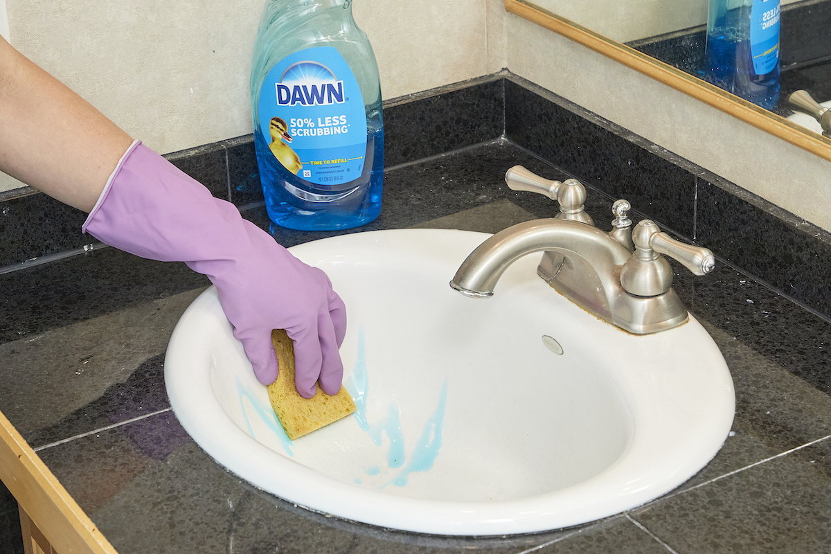 Woman wearing cleaning gloves cleans a bathroom sink with dish soap and a sponge.