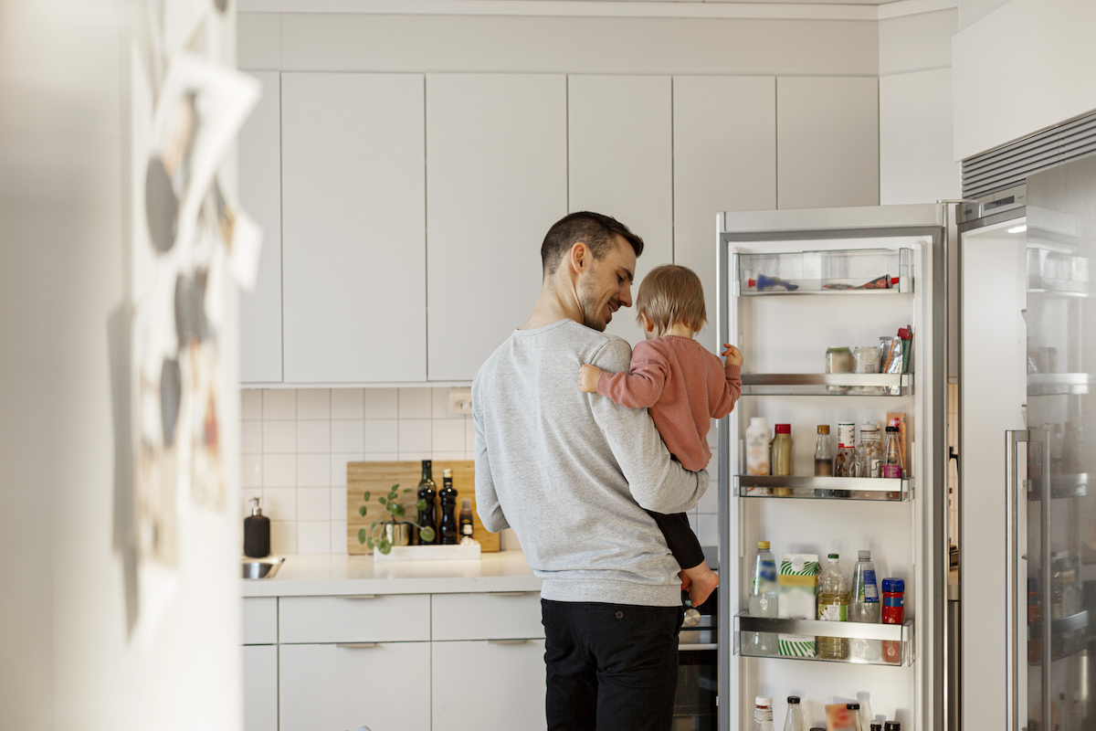 A man carrying a toddler whose hand is on the open refrigerator door.