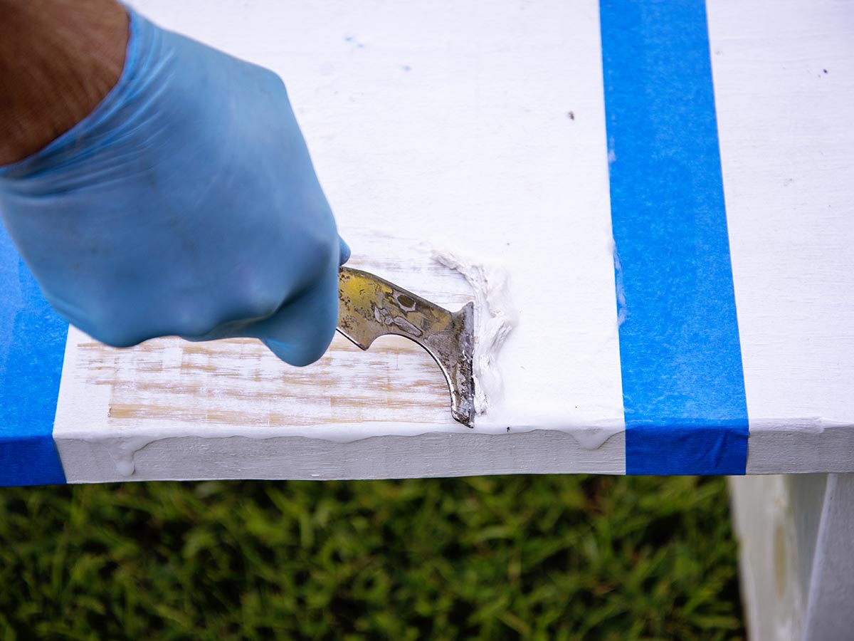 A gloved hand using a scraping tool to strip paint from a table