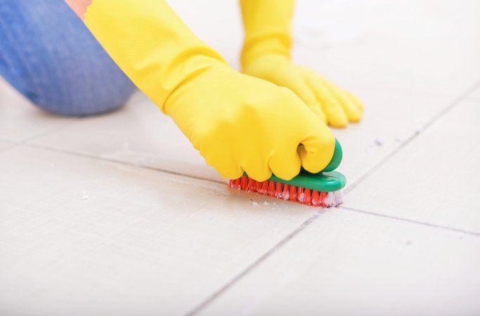 Person wearing yellow gloves cleans the joints of the floor tiles with a brush and powder.