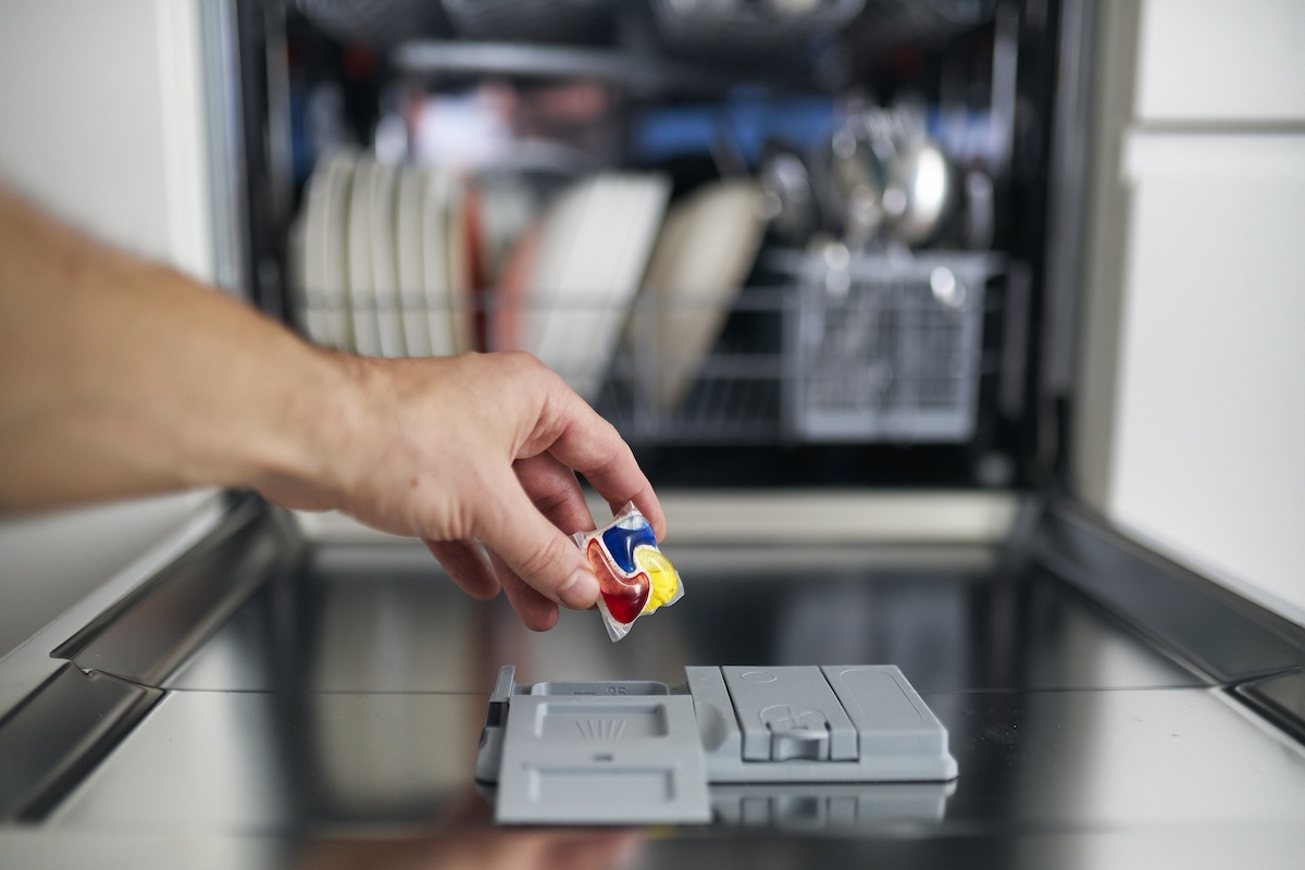 Person's hand inserting a red, blue, and yellow dishwasher pod into open dishwasher.