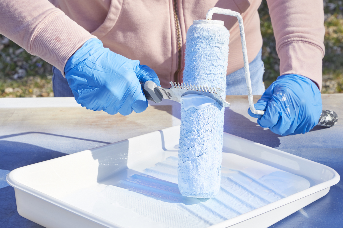 Woman uses a tool to clean paint off a paint roller that's resting in a paint tray.