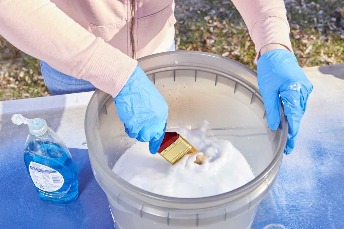 Woman washes a paint brush in a bucket of soapy water.