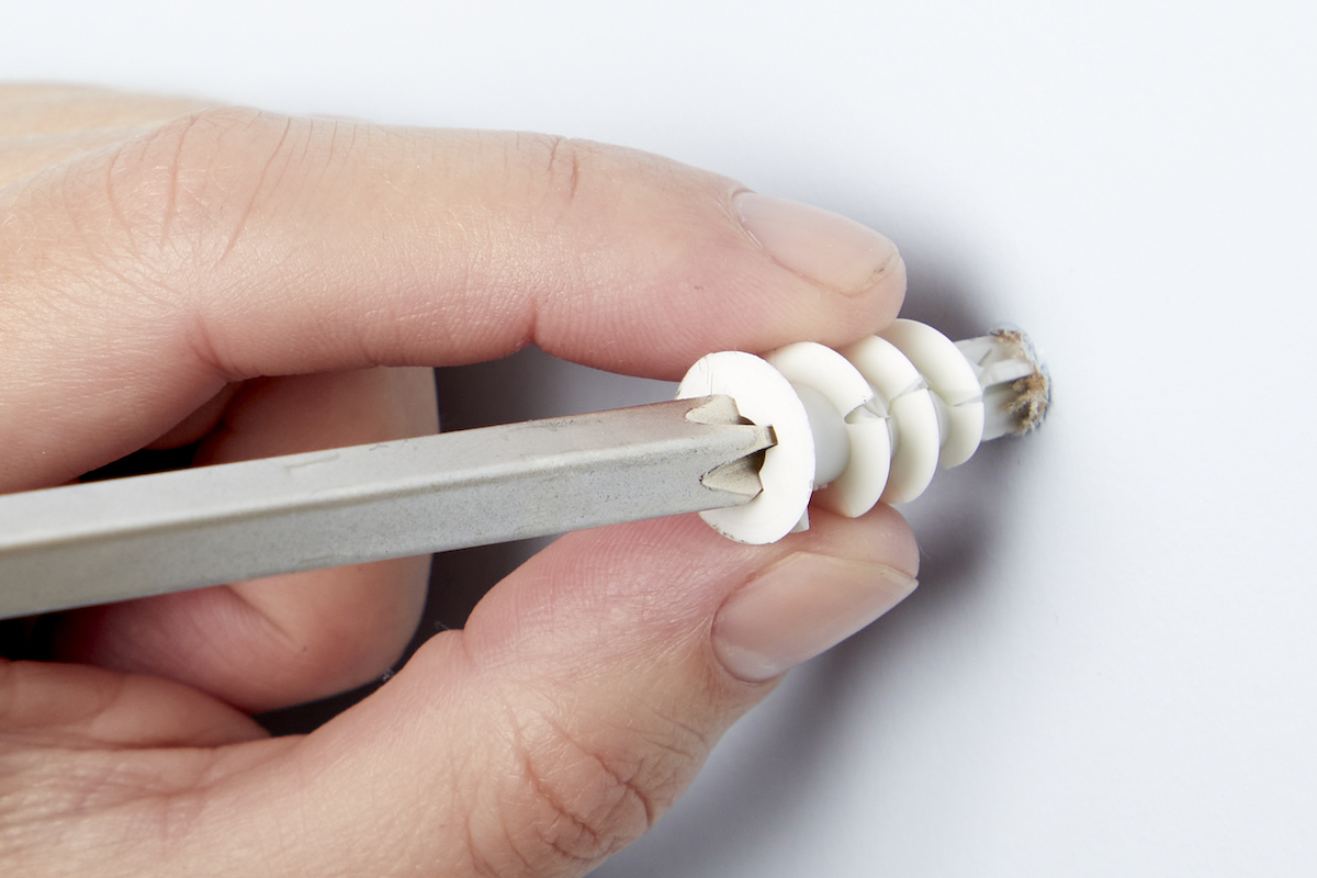 Close up view of a woman holding a drywall anchor between her fingers, with a screwdriver in the anchor.