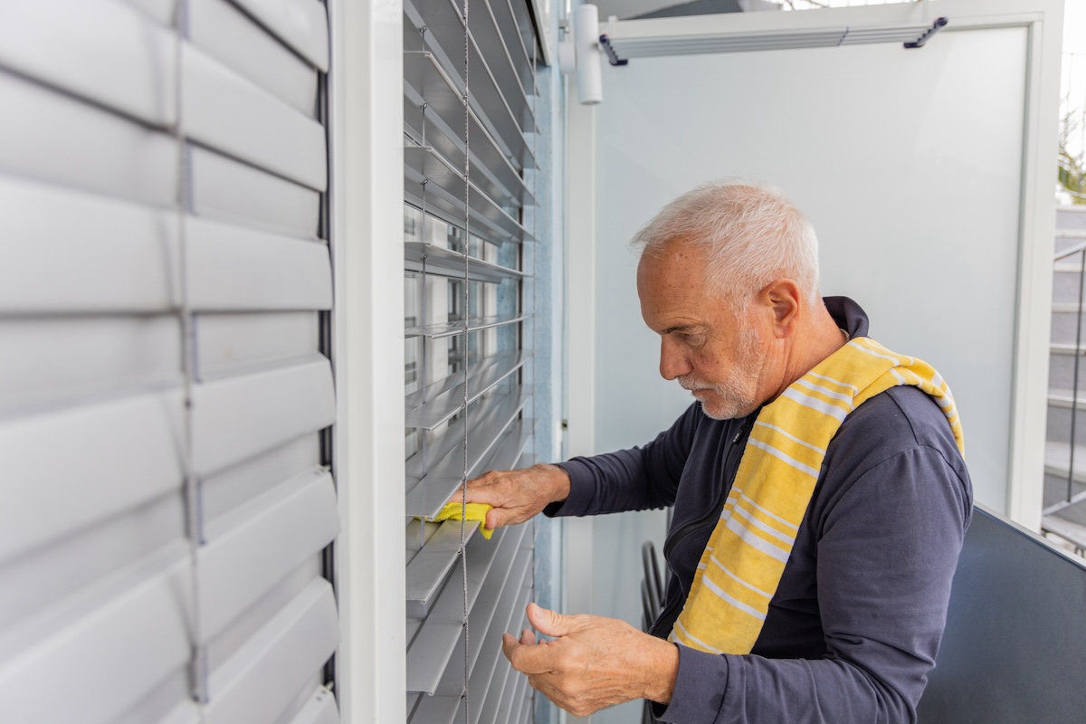 An older gentleman uses a yellow rag to clean outdoor blinds.