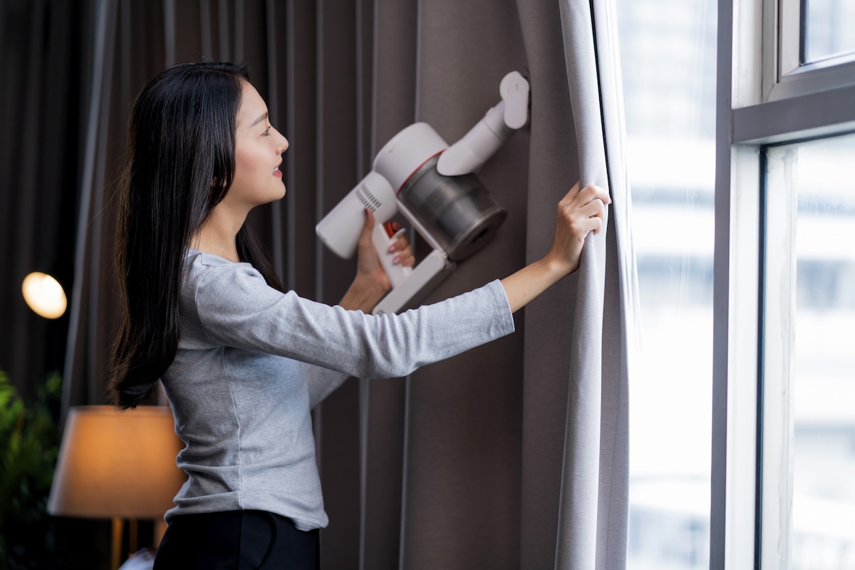 A woman wearing a grey shirt uses a handheld vacuum cleaner to clean dark curtains.