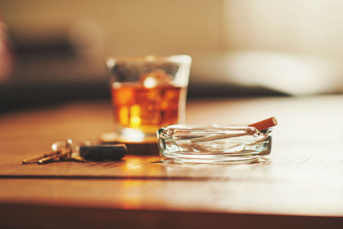 A glass of amber liquor and a cigarette in an ashtray sit atop a a wooden table.