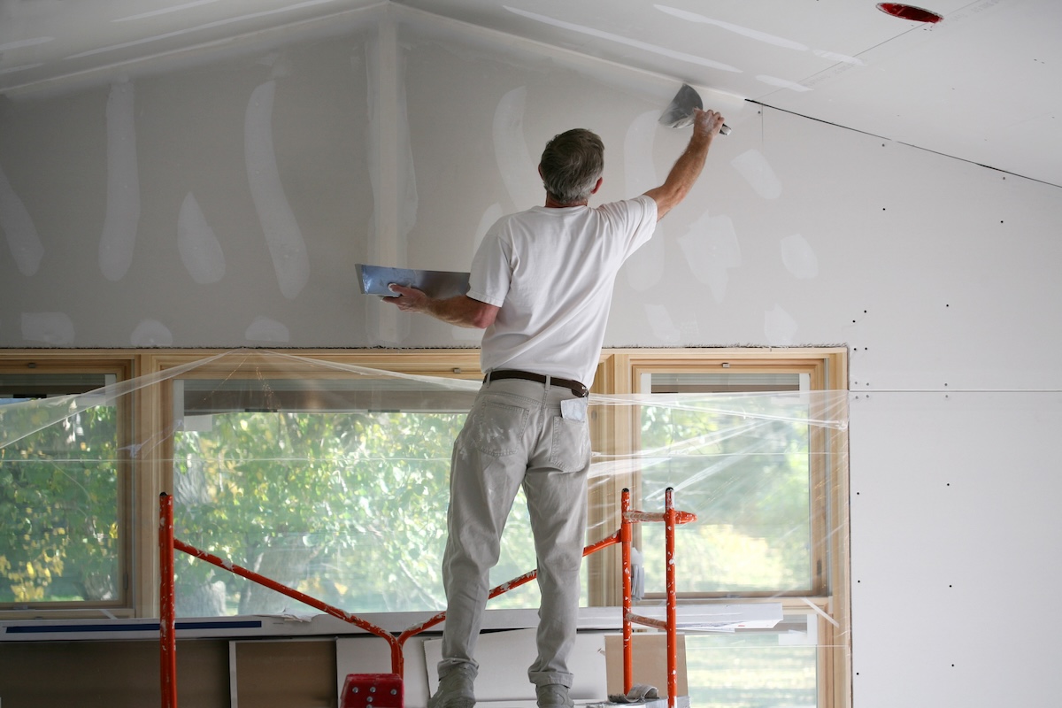 A man stands on scaffolding to apply spackle to an interior wall.