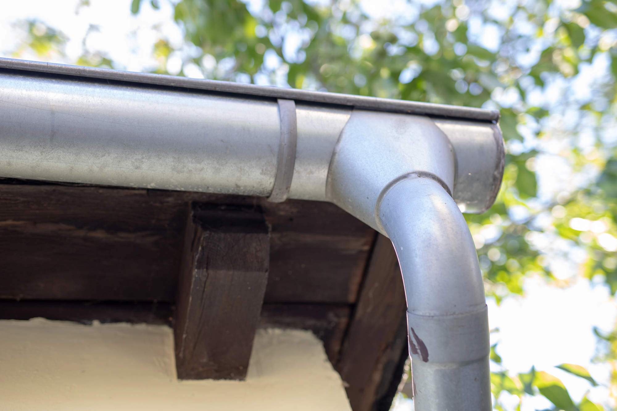 A sturdy zinc gutter and downspout hang over the dark-brown eaves of an old home.