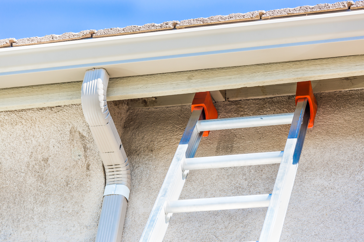 A ladder is leaning against a wall on a house with freshly installed white seamless gutters.