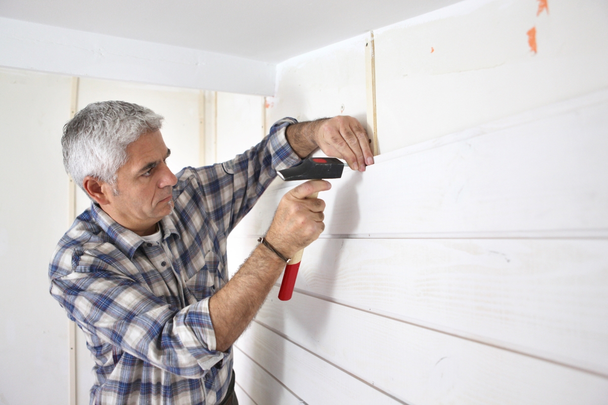 A grey-haired man is installing paneling on the wall with a hammer.